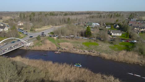aerial drone footage of people kayaking and paddle boarding with canoes and rowing on dark brown murky muddy river water underneath a bridge during a sunny day at võhandu marathon. spectators on shore