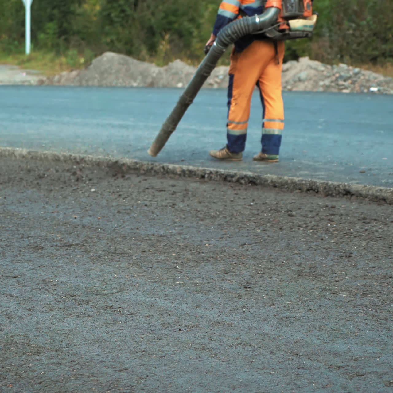 Worker in protective clothes blows out the pieces of old asphalt from the pits at road construction site. Road works concept. Worker is using compressed air to blow off dust on the road.