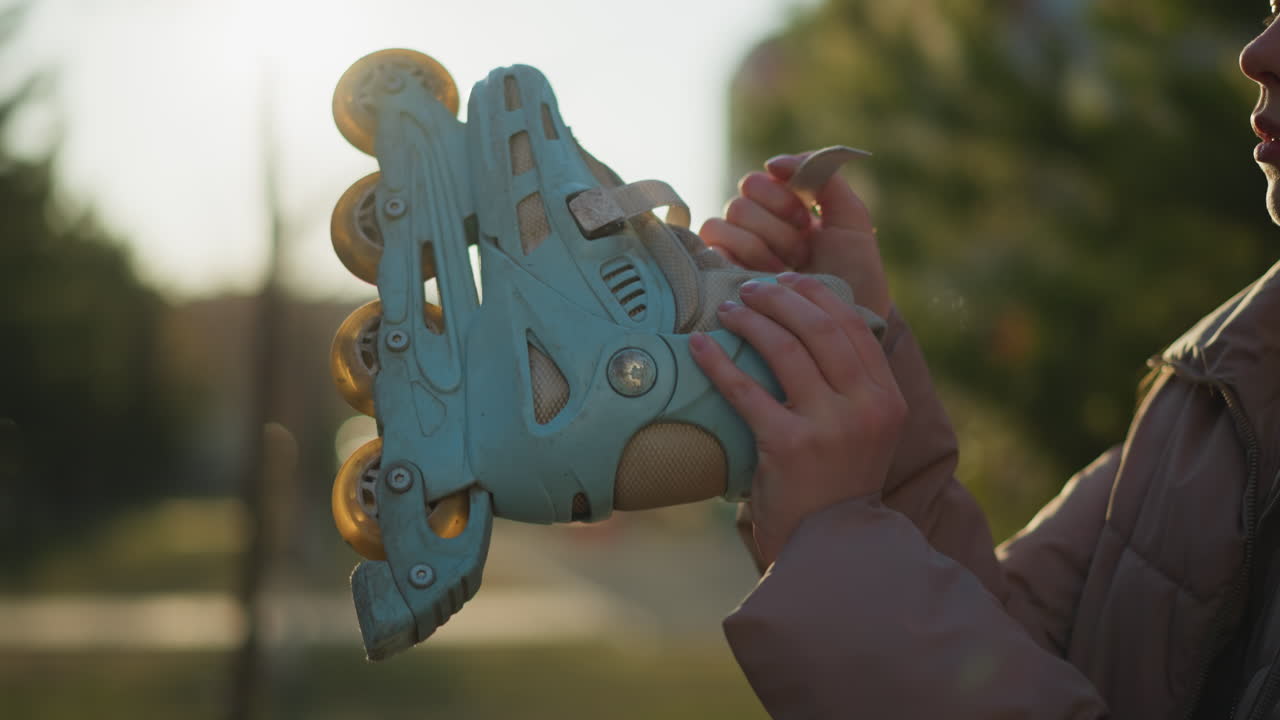 A young woman wearing a peach jacket is partially visible, with only half of her face showing, as she carefully adjusts the straps of her light blue rollerblades in a sunlit park