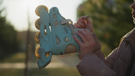 a young woman wearing a peach jacket is partially visible, with only half of her face showing, as she carefully adjusts the straps of her light blue rollerblades in a sunlit park