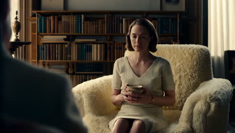 woman sitting in a fluffy armchair with a bowl, facing another person in a room with a bookshelf
