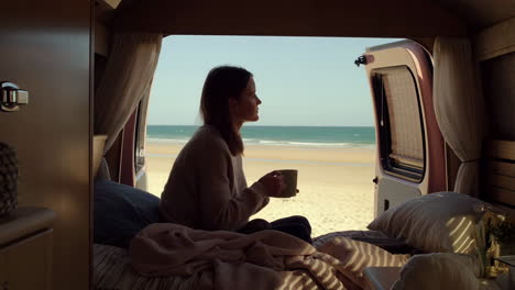 woman enjoying a drink with a beach view from inside a camper van