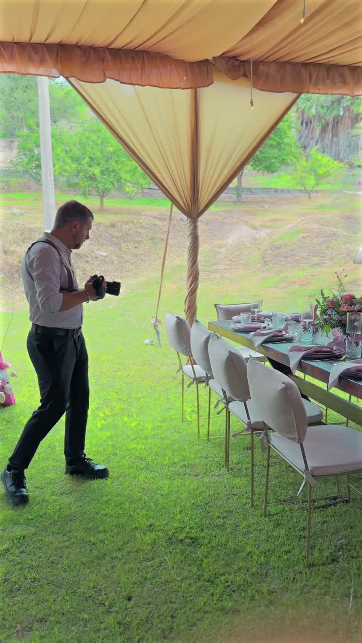 Photographer with camera walks on grass under beige tent, photographing decorated baptism table setting