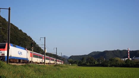 railway train passing by a agricultural land with mountains and forest in the background