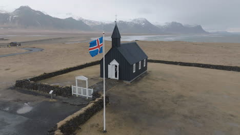 Drone-Shot-of-Black-Church-and-National-Flag-of-Iceland,-Famous-Budakirkja-on-Snaefellsnes-Peninsula