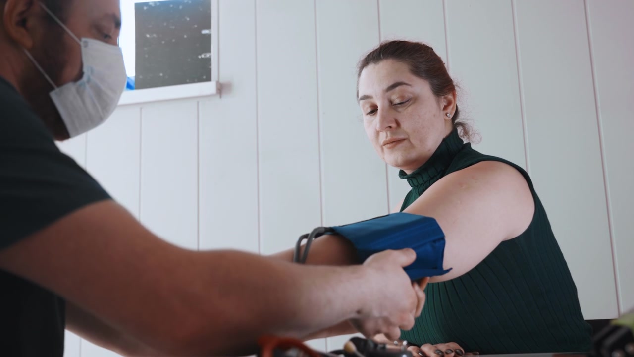 Mulher branca em visita domiciliar como enfermeira comunitária aplica manguito de pressão arterial enquanto médico mascarado auxilia, interior aconchegante com luz natural suave, atendimento atencioso à beira do leito durante monitoramento de saúde de rotina.