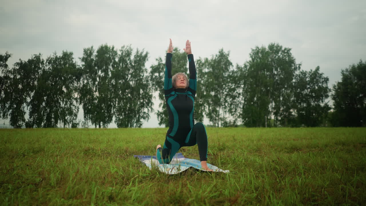Middle-aged woman in green and black suit outdoor in vast grassy field on yoga mat practicing low lunge pose with arms wide open, under cloudy sky and trees in background