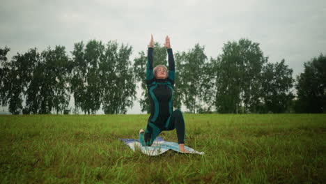 middle-aged woman in green and black suit outdoor in vast grassy field on yoga mat practicing low lunge pose with arms wide open, under cloudy sky and trees in background