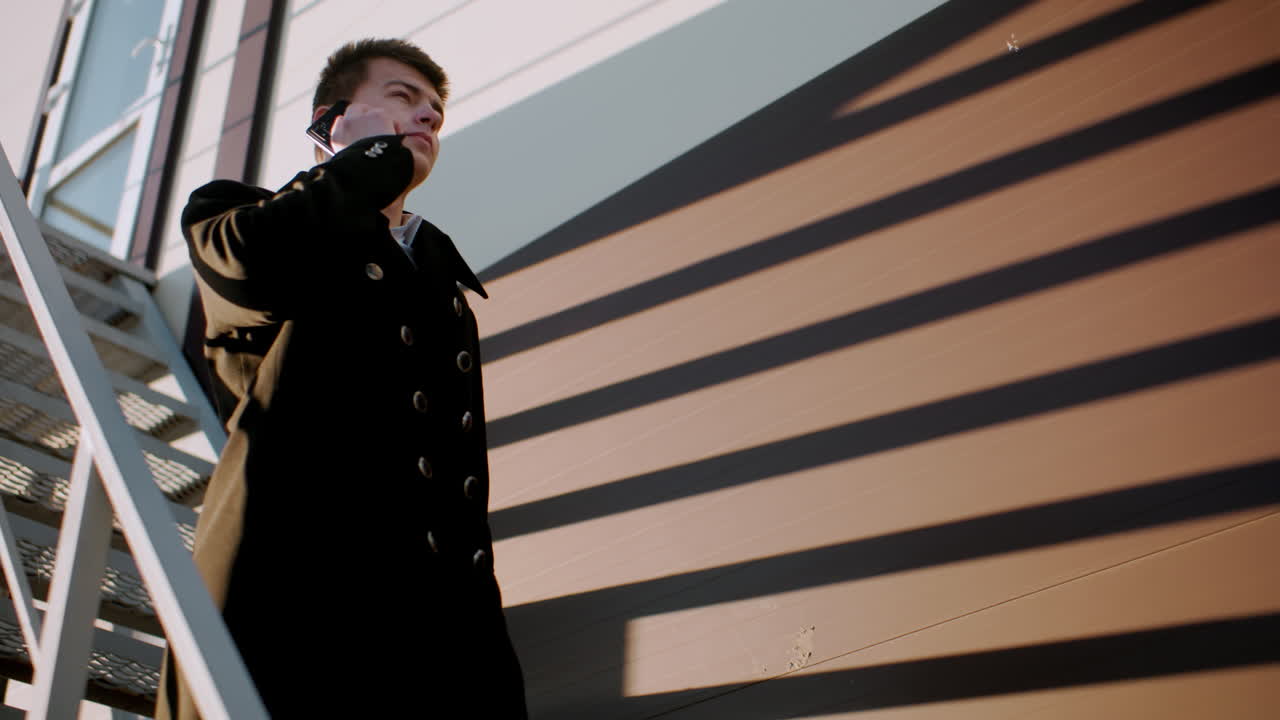 검은 코트 를 입은 사업가 가 전화 를 하는 동안 계단 을 내려가고, 서류 가방 을 들고 있으며, 건물 의 유리 창문 을 배경 으로 반사 하고 있다