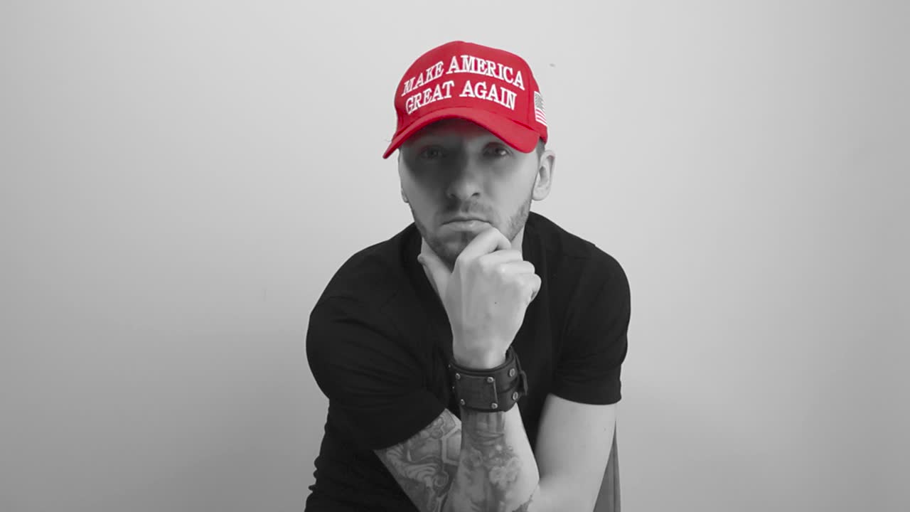 Grayscale footage of a tattooed man sitting and putting on a red colored MAGA Make America Great Again hat, then puts his hand on his jaw showing he is thinking and watches directly forward.