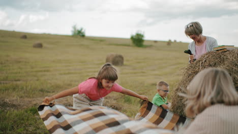 close-up rear view of mother and daughter spreading cloth on ground while woman holds dog leash, nearby, a little child watches, creating a warm, peaceful family moment in the open countryside