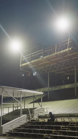 a man dressed in black and wearing a jacket sits quietly in an empty stadium at night, with a soccer ball by his side, the background is illuminated