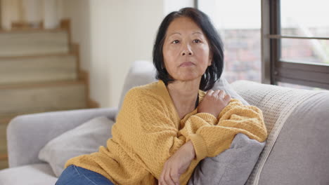 senior asian woman in yellow sweater sitting on couch, looking thoughtful at home