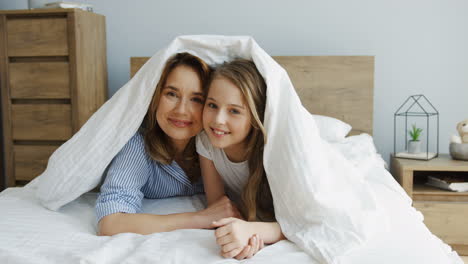 bonita madre caucásica y su linda hija sonriendo y mirando fuera de la manta mientras se acuesta en la cama por la mañana