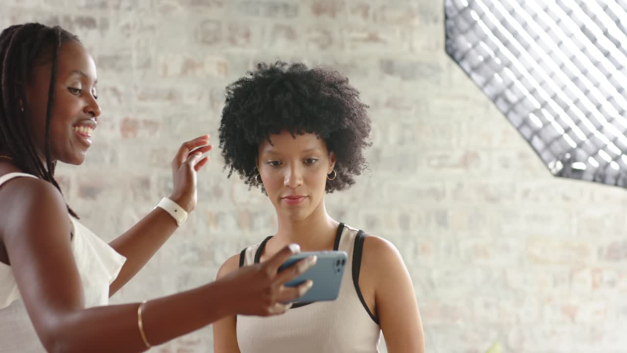 African American women friends calling, showing smartphone for selfie and hugging near softbox