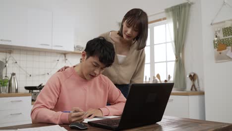 frustrated asian man managing family finances and doing research on computer at dining table. his wife walking over to cheer him up while he is writing in notebook