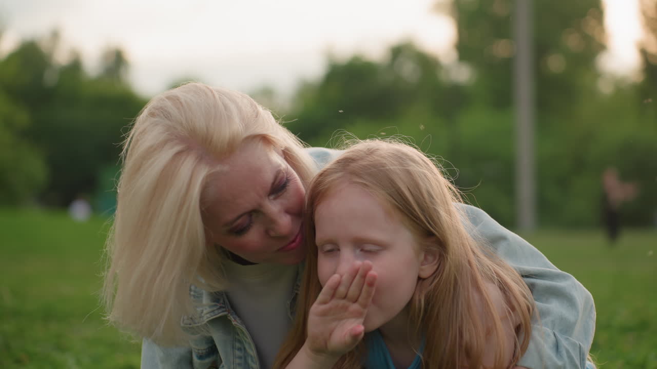 young girl seated on grass rubbing saliva on back of hand then face while mother hugs from behind in park, playful caring moment with blurred people and trees in background warm light