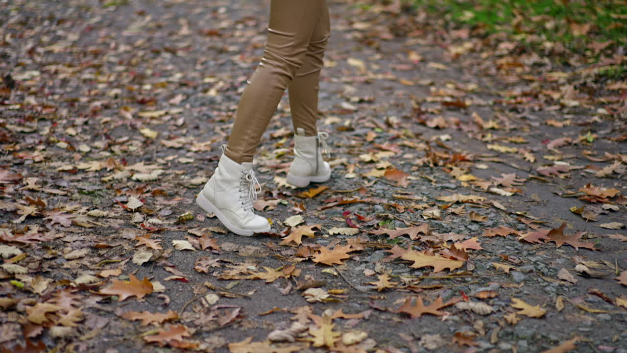 Unrecognized lady in beige pants and white jacket and boots walking in the park. Long-haired brunette turning around demonstrating her look.