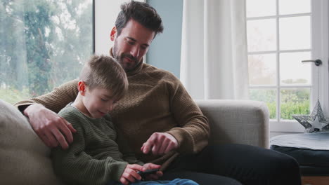 Padre-Jugando-Videojuegos-Con-Su-Hijo-En-El-Teléfono-Móvil-Sentado-En-El-Sofá-En-Casa