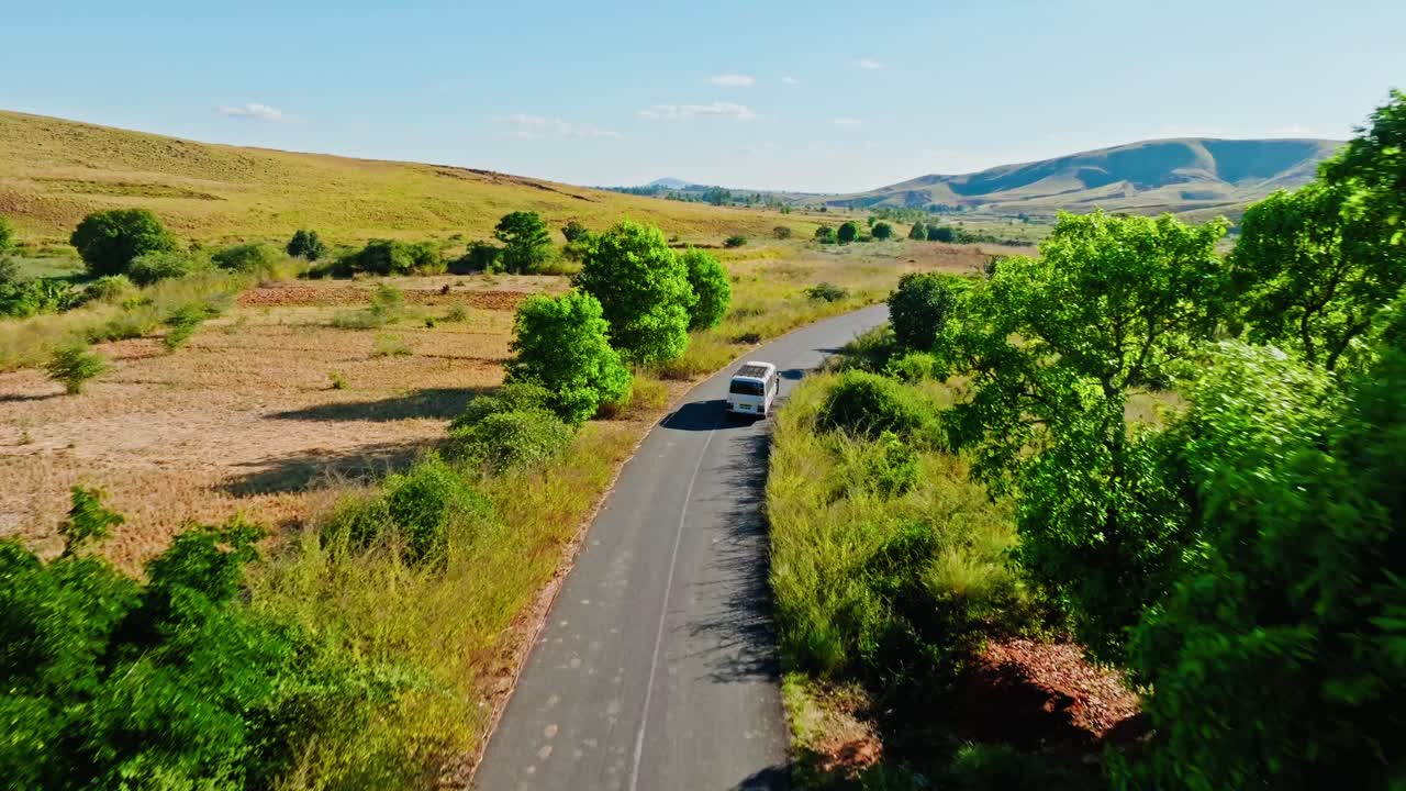 Aerial view of a white bus driving along a remote road through grassy highlands and green trees in Madagascar. Peaceful rural transportation scene under a clear sky.