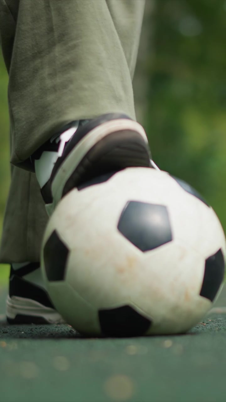 Ayak, yıpranmış futbol topunun sabit konumuna biraz daha yaklaşıyor. Yanında görünen kirli, yıpranmış siyah beyaz spor ayakkabılar. İnce detaylar arasında kişinin üzerindeki haki pantolon yer alıyor.