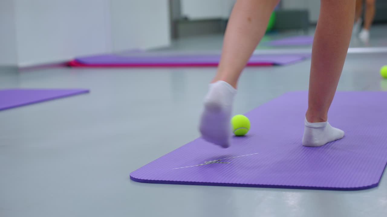 vista de la pierna trasera en calcetines moviéndose hacia adelante y hacia atrás en una alfombra púrpura con una pelota de tenis verde, el espejo del gimnasio refleja el fondo, capturando la sesión de fitness y el movimiento de los pies durante el ejercicio de coordinación