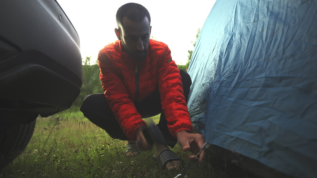red jacket camper trekker hammering tent pegs setting up the campground in green meadow grass