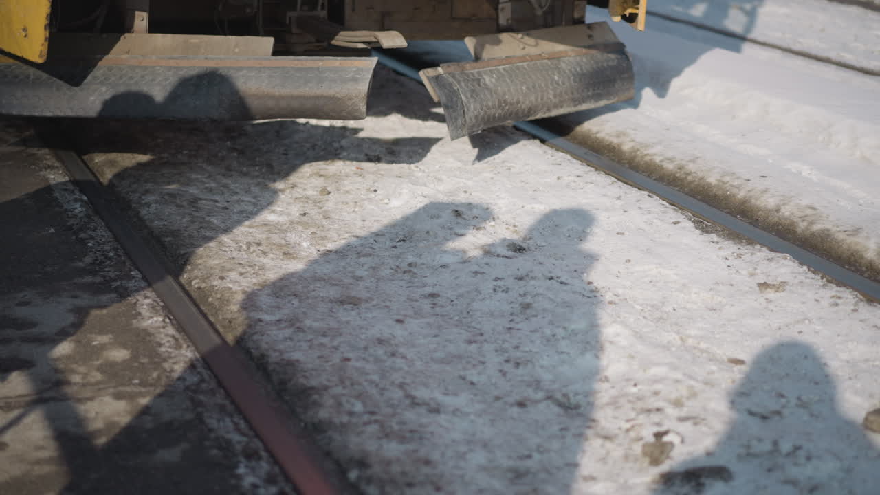 Winter street scene with shadows of people across snowy rails as tram passes, crisp sunlight on gritty ice, textured ground, quiet urban mood, low perspective at track edge showing motion, contrast