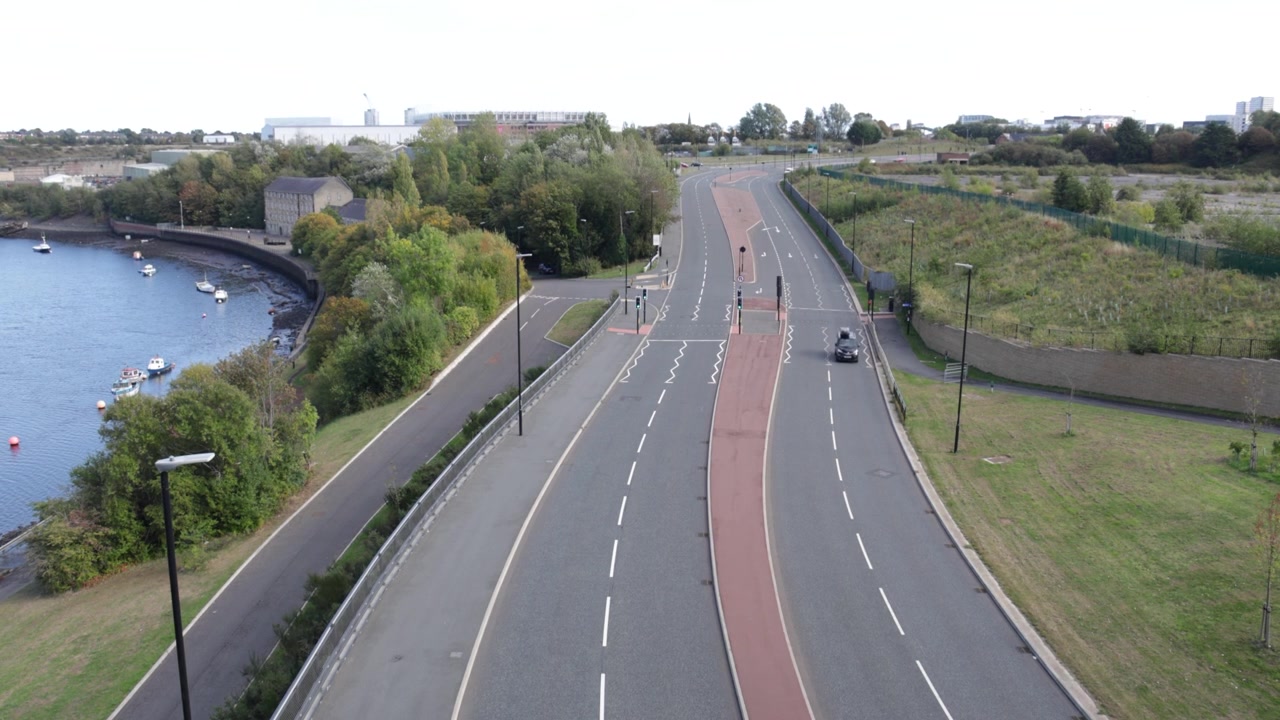 Ripresa dall'alto di una strada asfaltata a più corsie vuota che costeggia un fiume calmo con barche. Mostra l'infrastruttura urbana e la pianificazione urbana sostenibile con spazi verdi