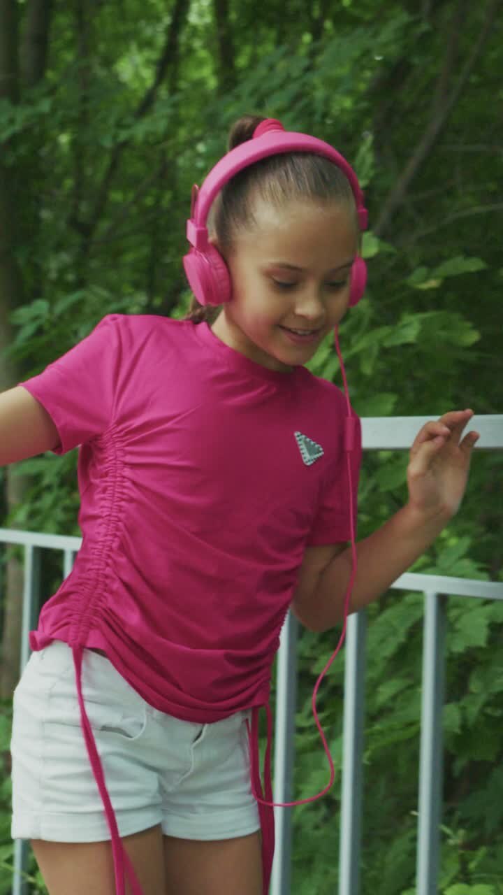 A young girl enjoys her favorite music while wearing vibrant pink headphones and a stylish top, expressing joy and rhythm in a lush green setting
