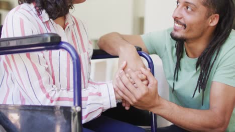 Happy-biracial-woman-in-wheelchair-and-smiling-male-partner-holding-hands-and-talking-in-living-room