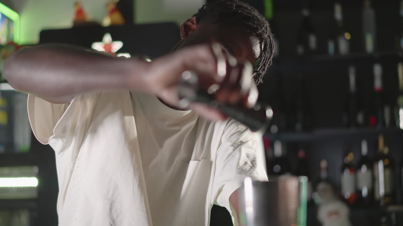 Bartender pouring liquor into jigger at bar, preparing cocktail with focus on measuring, bartender in white shirt with blurred background of neon lights, bar shelf, and liquor bottles