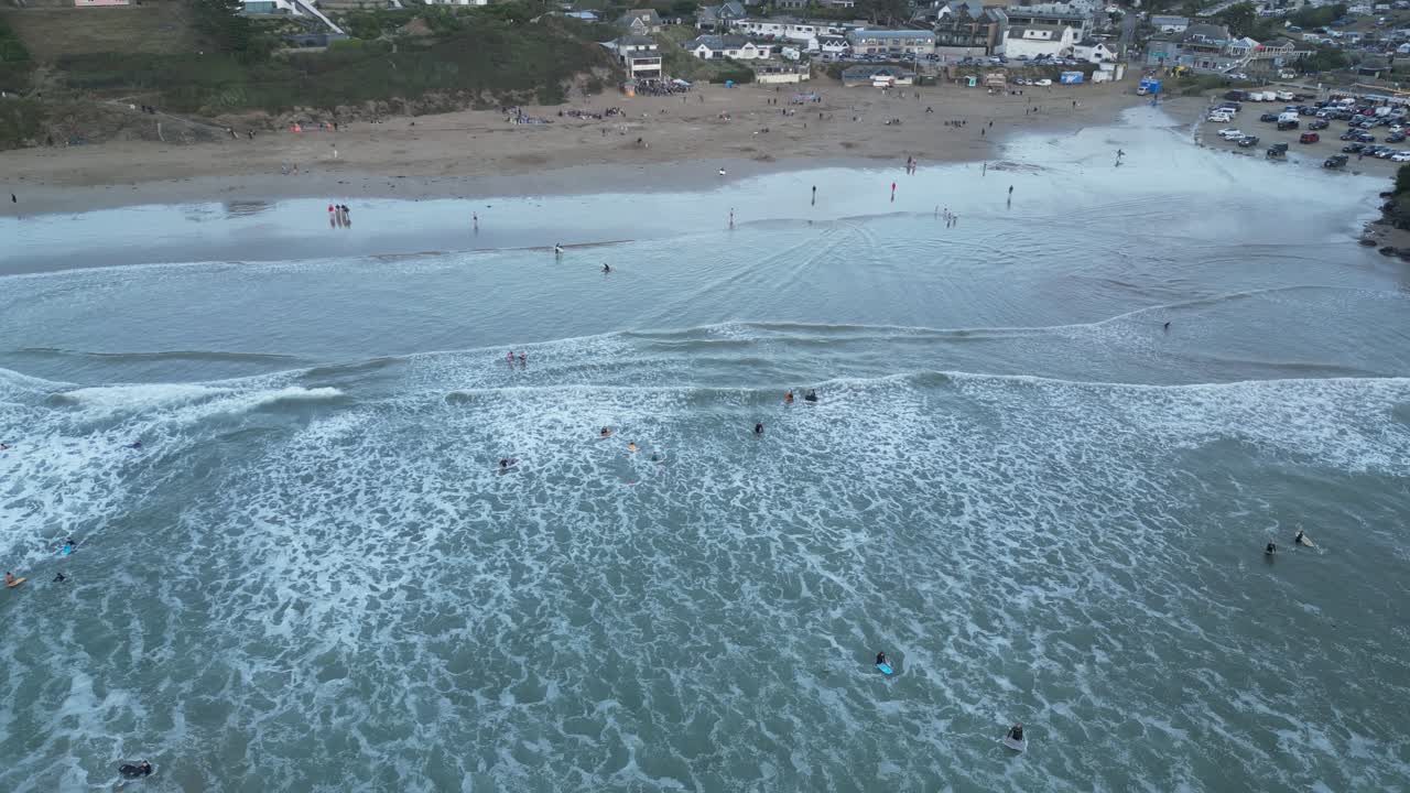 Stunning drone video captures Polzeath Beach's dynamic waves with surfers braving the waters, under soft evening light. The sweeping aerial shot includes sandy shores and beachfront buildings
