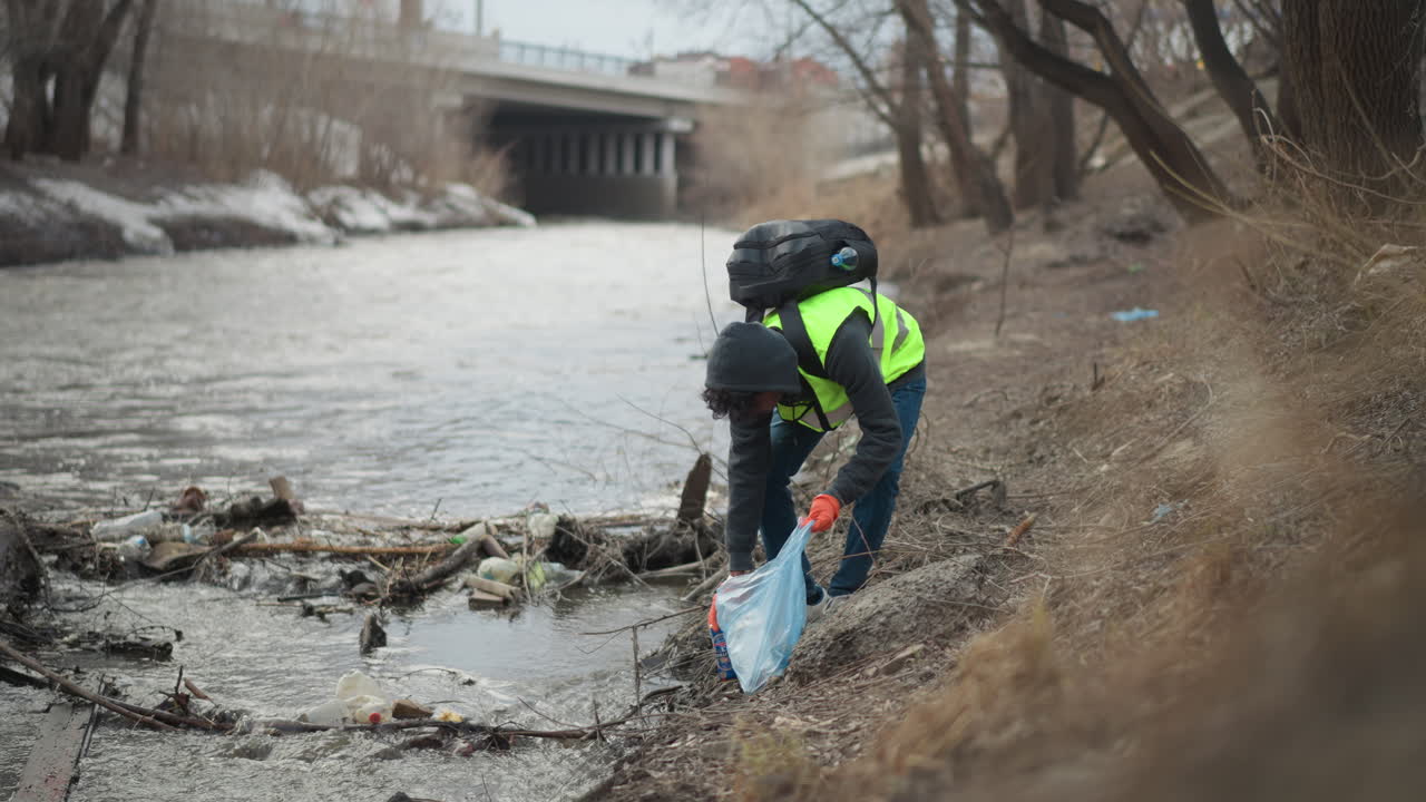 Volontario con giubbotto riflettente e guanti raccoglie una bottiglia di plastica dalla sponda inquinata del fiume, mettendo i rifiuti in un sacco blu durante uno sforzo di pulizia ambientale per proteggere la natura.