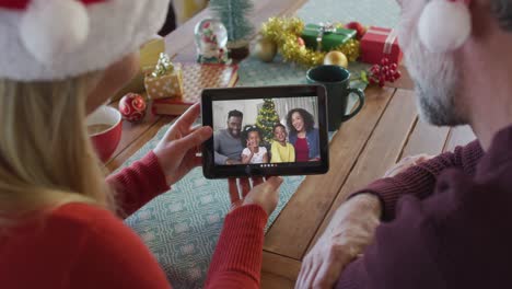 Lächelndes-Kaukasisches-Paar-Mit-Weihnachtsmützen,-Das-Ein-Tablet-Für-Einen-Weihnachtsvideoanruf-Mit-Der-Familie-Auf-Dem-Bildschirm-Nutzt