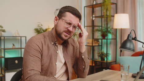tired ill caucasian man using laptop and suffering from headache problem tension sitting at table