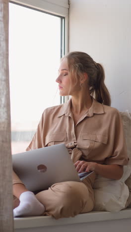 young woman gets distance education via modern laptop sitting near window behind brown curtain in comfortable room at home slow motion
