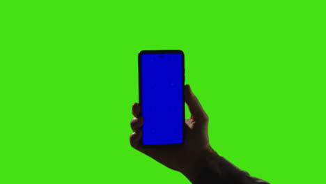 close up of man holding blue screen mobile phone against green screen background with low key lighting