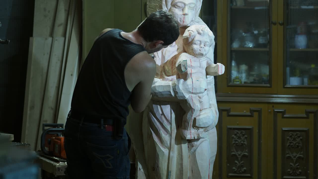 An artisan in a sleeveless shirt meticulously sculpts the arms of a wooden Virgin Mary and Child statue, focusing on delicate details in a traditional workshop setting