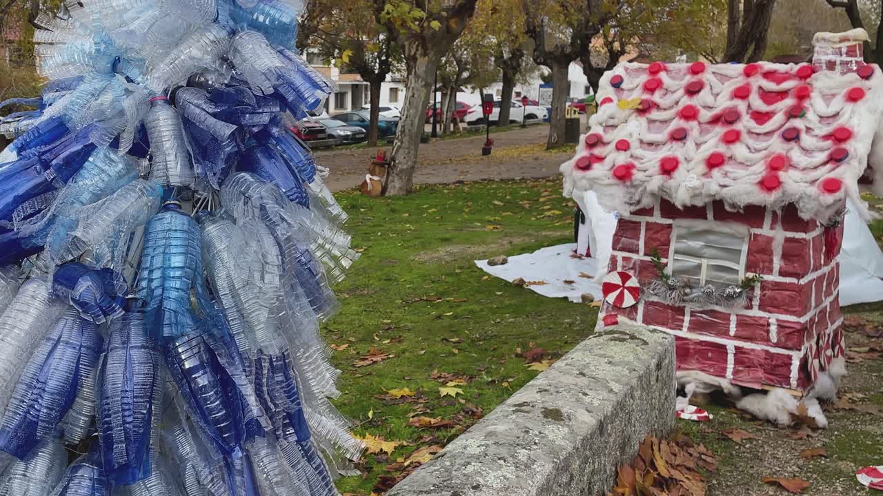 Tomada panorâmica lateral em câmera lenta em um parque infantil mostrando uma árvore de Natal feita com tiras de garrafas plásticas recicladas. Uma cabana de Natal vermelha construída com materiais reciclados como lã e plástico.
