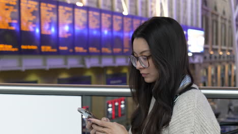 young woman texting on her smartphone while waiting for her train, timetable boards can be seen in the background