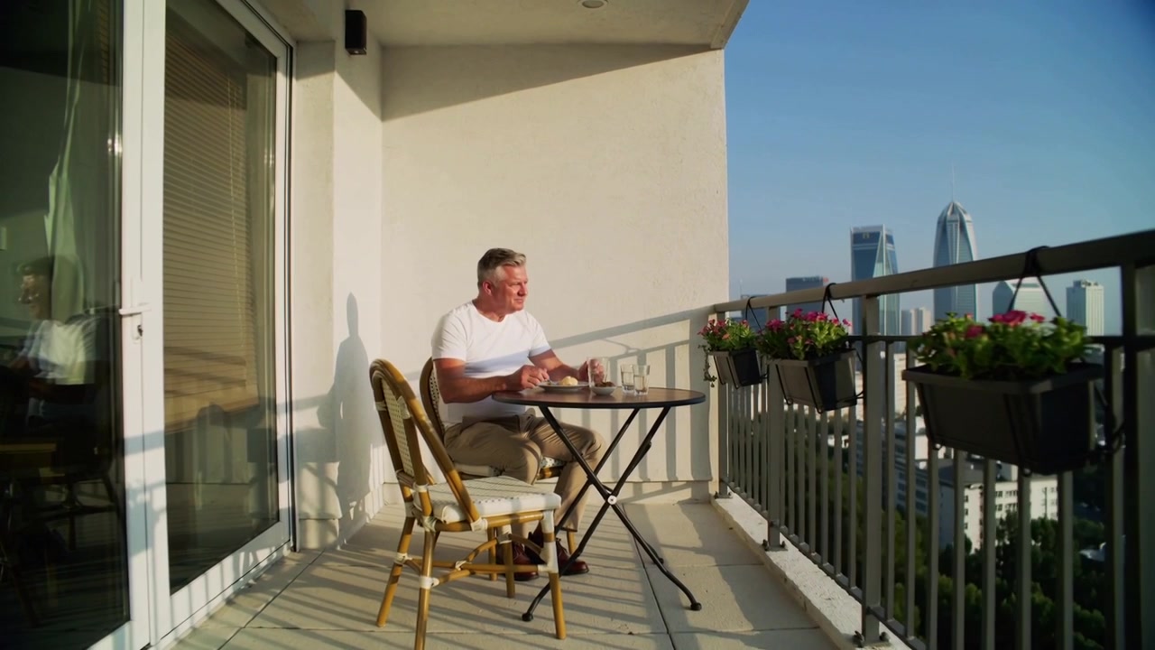 Notando la luce del sole, un uomo con una maglietta bianca sorseggia da una tazza, maneggia le posate e guarda lo skyline dal balcone.