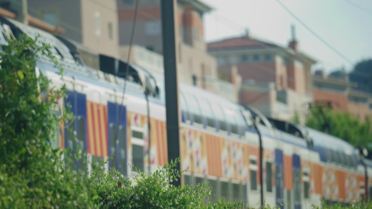 Treno che passa dietro cespugli verdi, con grafiche esterne colorate e uno sfondo urbano sfocato e delicato.
