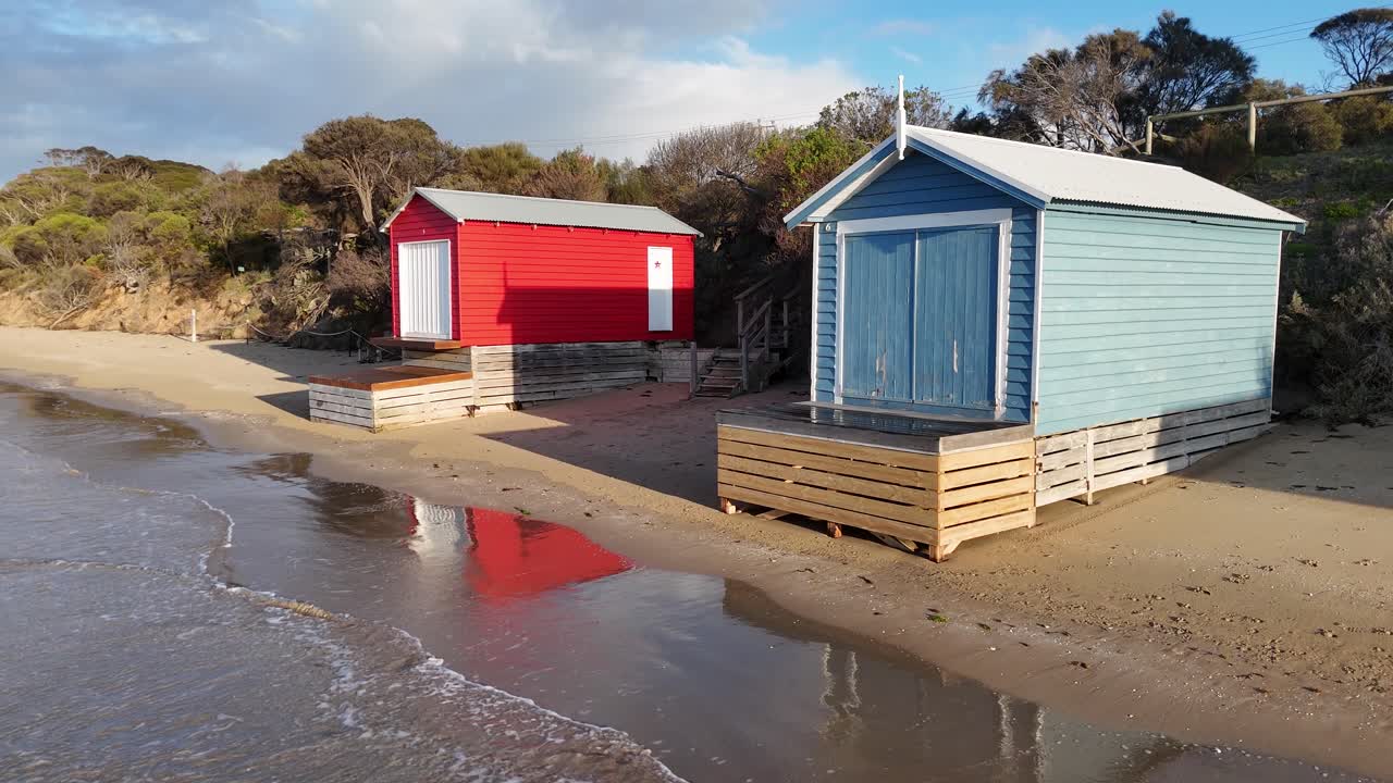 Aerial camera glides past vibrant beach huts on sandy shore, morning sunlight, gentle waves visible