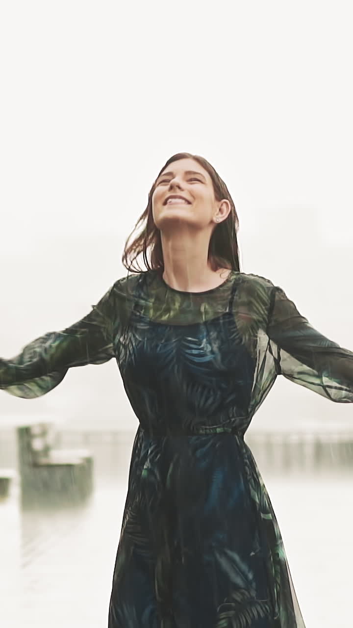 Positive brunette woman in elegant wet long dress stands spinning under heavy rain on viewing deck against blurry city in mist on summer day slow motion
