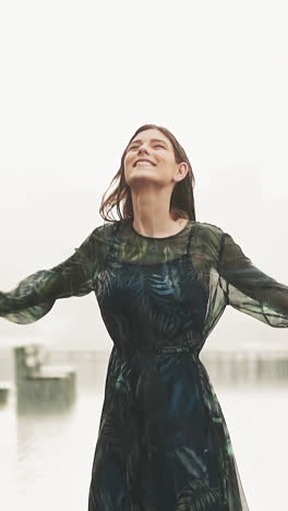 positive brunette woman in elegant wet long dress stands spinning under heavy rain on viewing deck against blurry city in mist on summer day slow motion