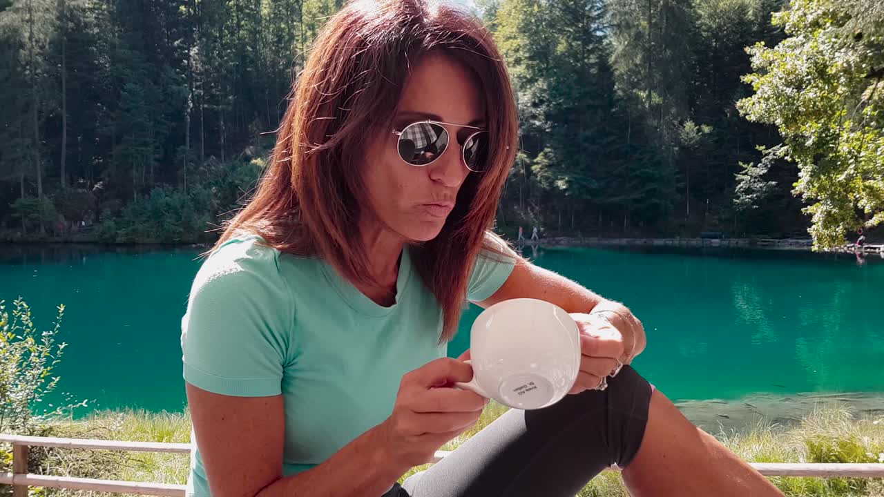 mujer mayor deportiva y atractiva tomando un café, hablando y relajándose junto a un lago en la naturaleza después de hacer ejercicio en verano