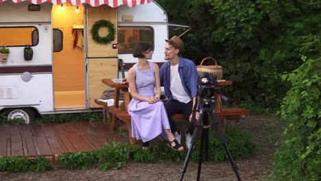 Popular-Bloggers-Couple-Greeting-Their-Followers,-Recording-Video-While-Sitting-At-The-Wooden-Table-In-Front-The-Wheels-House,-Talking-And-Looking-At-Camera-On-Tripod-1