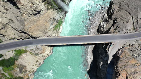 Tiro-De-Dron-Del-Río-De-Agua-Azul-Turquesa-Que-Fluye-Bajo-Un-Puente-En-La-Cordillera-De-Karakoram-A-Lo-Largo-De-La-Carretera-De-Karakoram,-Toma-Aérea-Ascendente