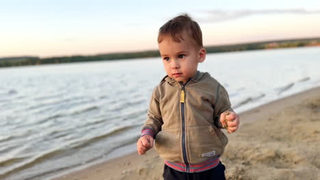 little cute toddler boy plays in the sand at shore. kid takes some sand with bare hands and throws it to the grass.